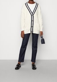 Cárdigan de punto en crema con ribetes y botones en azul marino, combinado con pantalones de mezclilla oscuros y mocasines negros, sostenido con un pequeño bolso negro.