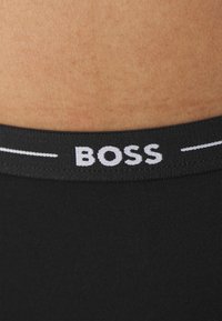 Ceinture noire avec le logo "BOSS" en blanc, fabriquée dans un tissu élastique et lisse. L'arrière-plan présente une texture de teint de peau.