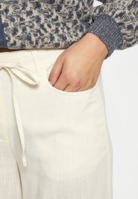 Bež lanene hlače z sprednjim vezanjem in stranskimi žepi, v kombinaciji z teksturiranim puloverjem v modri in kremni barvi.