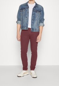 Veste en denim bleu clair avec deux poches frontales, t-shirt blanc, pantalon slim bordeaux et baskets blanches avec accents beiges.