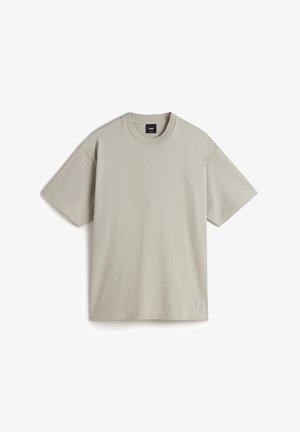 T-shirt à manches courtes gris clair en coton. Doté d'un col rond et d'un petit patch logo en bas à gauche. Texture douce.
