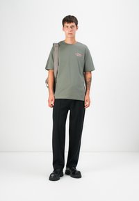 Olivengrünes T-Shirt mit kurzen Ärmeln und Logodetail, kombiniert mit schwarzen Hosen und klobigen schwarzen Schuhen, Modell hält einen grauen Rucksack.