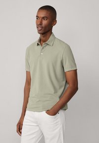 Polo shirt verde chiaro con colletto e due bottoni, realizzato in tessuto morbido. Indossato con pantaloni bianchi, design minimalista, maniche corte.