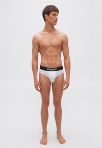 Jeune homme debout pieds nus portant un slip blanc avec une ceinture noire et le logo "VERSACE", regardant vers sa gauche sur un fond uni.