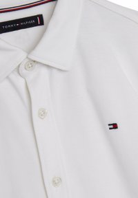 Polo shirt bianco in cotone con maniche corte, tre bottoni e un piccolo logo ricamato in blu navy, rosso e bianco sul lato sinistro del petto.