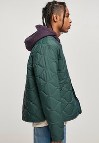 Chaqueta verde acolchada con un patrón ondulado, bolsillos laterales y una sudadera morada debajo, mostrada desde un ángulo lateral.