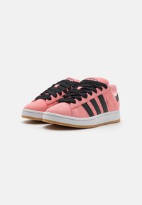 Růžové semišové tenisky mají černé tkaničky a tři černé proužky na boku, s bílou mezipodešví a gumovou podrážkou.