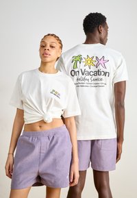 Weißes T-Shirt mit Knoten und buntem Grafikdesign; kombiniert mit helllila Cordshorts mit elastischem Bund und Seitentaschen.