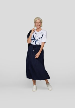 Weißes T-Shirt mit blauem abstraktem Design, kombiniert mit einem langen, marineblauen Faltenrock. Modell trägt weiße Sportschuhe und hält eine schwarze Jacke.