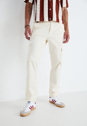 Pantalones cargo beige con bolsillos laterales, hechos de tejido ligero. Combinados con una camisa de manga corta estampada y zapatillas blancas con detalles rojos.