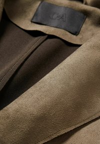 Tejido de ante marrón con una textura suave, que presenta una etiqueta negra. El diseño incluye un collar y detalles de costura visibles.