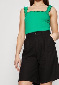 Femme portant un débardeur froncé vert vif avec des bretelles volantes et un short noir ample taille haute avec des poches.