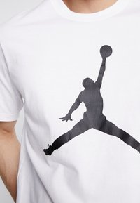 Hvid bomulds t-shirt med en stor sort silhuet grafik af en person, der hopper for at lave et basketballskud. Rund halsudskæring, kortærmede.