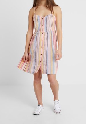 Robe A-line rayée avec des bretelles spaghetti, boutons sur le devant, dans des teintes pastel de rose, bleu et jaune. Portée avec des baskets blanches.