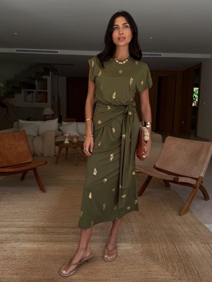 Robe midi vert olive avec des motifs en feuilles d'or, manches courtes et taille nouée. Portée avec des sandales transparentes, mettant en valeur une texture lisse.