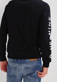 Svart sweatshirt med ribbad halsringning och muddar, med vit text längs ärmen. Kombinerad med blå denimjeans med fickor.