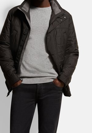 Man draagt een zwarte doorgestikte jas over een lichtgrijze trui en donkere spijkerbroek, handen in de zakken, tegen een effen lichte achtergrond.