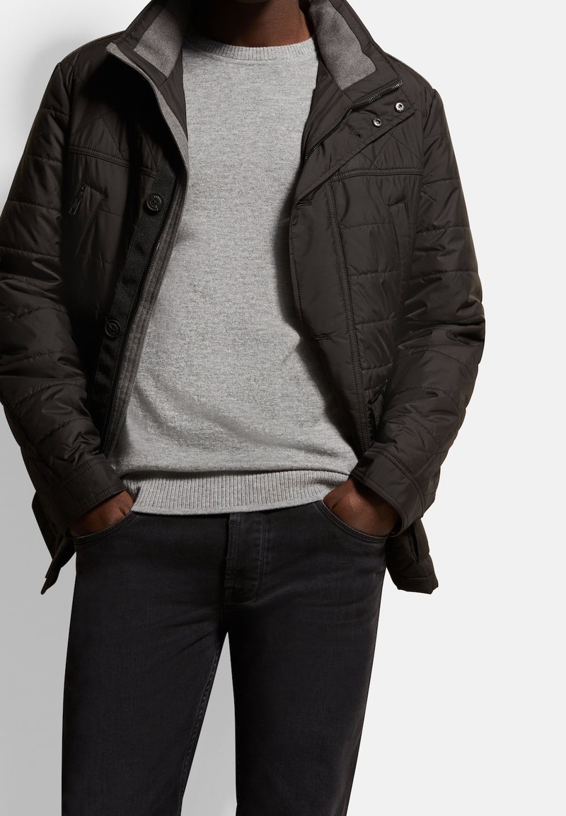 Homme portant une veste matelassée noire par-dessus un pull gris clair et un jean foncé, les mains dans les poches, sur un fond clair uni.