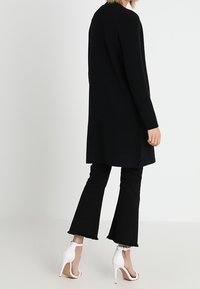 Svart lång kofta med rak skärning, mjuk textur. Kombinerad med svarta utsvängda byxor och vita klacksandaler, som visar upp en modern outfit.