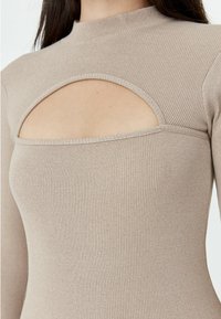 Top beige à manches longues côtelé, mettant en avant un col rond et un design découpé au niveau de la poitrine, soulignant une texture lisse et une coupe ajustée.