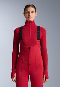 Piros sportos body bordázott garbónyakkal és fekete állítható pántokkal. Elöl cipzárral és testhez simuló, sima textúrájú sziluettel rendelkezik.