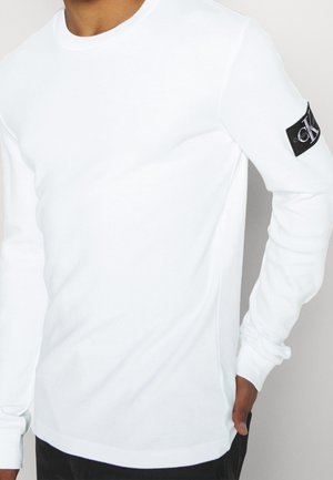Mężczyzna noszący gładką białą koszulę z długim rękawem z czarną naszywką z logo Calvin Klein na górnym lewym rękawie, ręka w kieszeni.