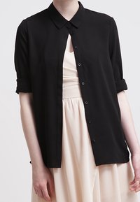 Chemise noire à manches courtes avec col et boutons, manches retroussées, tissu lisse et coupe droite, portée par-dessus une robe de couleur claire.