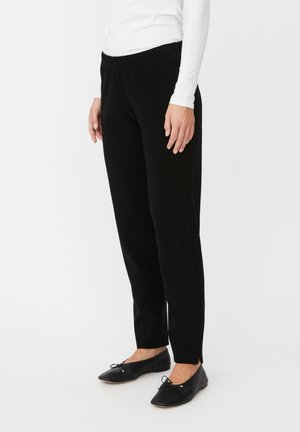 Femme portant un pantalon noir slim jusqu'à la cheville avec des fentes sur les côtés, associé à des ballerines noires et un haut blanc à manches longues.