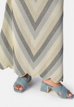 Pies con sandalias de tacón ancho de ante azul con punta abierta bajo una falda de punto con patrón de chevron y dobladillo festoneado en tonos beige y gris.