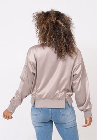 Lichtbeige bomberjack in satijnen afwerking, met een ribbelige zoom, ritszakken en een relaxte pasvorm. Geverfd over blauwe denimjeans.