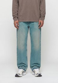 Άτομο που φοράει ξεθωριασμένο μπλε τζιν με ίσια πόδια, καφέ μακρυμάνικο πουκάμισο και λευκά αθλητικά παπούτσια, όρθιο μπροστά σε απλό φόντο.