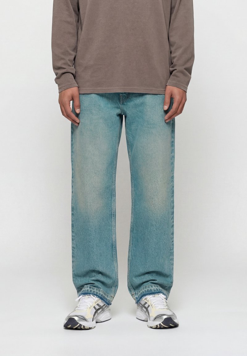 Άτομο που φοράει ξεθωριασμένο μπλε τζιν με ίσια πόδια, καφέ μακρυμάνικο πουκάμισο και λευκά αθλητικά παπούτσια, όρθιο μπροστά σε απλό φόντο.