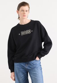 Felpa nera con scollo rotondo e vestibilità ampia. Presenta una scritta "BOSS" in bianco audace con stelle decorative. Abbinata a jeans blu.