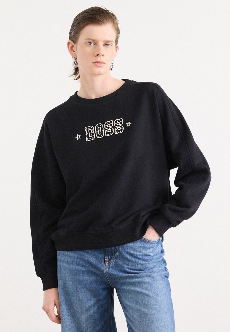 Felpa nera con scollo rotondo e vestibilità ampia. Presenta una scritta "BOSS" in bianco audace con stelle decorative. Abbinata a jeans blu.