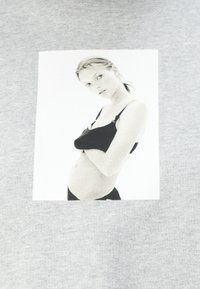 Fotografia in bianco e nero di una donna incinta con reggiseno e mutandine nere, che guarda la fotocamera con le braccia incrociate sul petto.