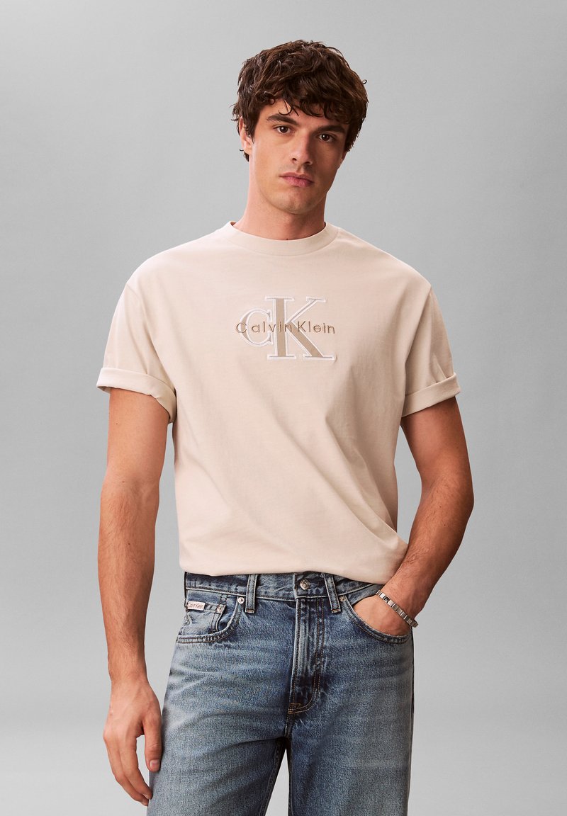 Ung mand med kort krøllet hår iført en beige Calvin Klein t-shirt og blå jeans, stående med den ene hånd i lommen mod en grå baggrund.