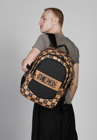 Mochila de checkered negra y naranja con un gráfico central, que presenta el texto "One Piece" y acentos de emblema, fabricada con tela duradera y correas ajustables.