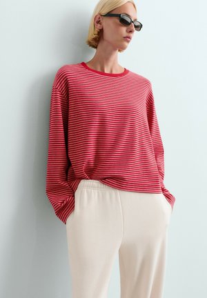 Femme portant de petites lunettes de soleil noires, un t-shirt à manches longues rayé rouge et blanc, et un pantalon beige clair, les mains dans les poches, devant un mur uni.