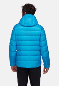 Giacca a vento blu con design trapuntato, cappuccio e logo bianco "MAMMUT" sulla schiena, caratterizzata da una texture liscia e lucida.