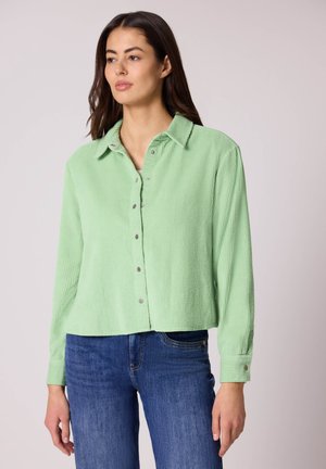 Donna che indossa una camicia di velluto a coste verde chiaro con bottoni e jeans blu, in piedi contro uno sfondo chiaro uniforme, con lo sguardo leggermente rivolto a sinistra.