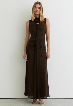 Vrouw met blond haar, gekleed in een mouwloze, tot op de vloer vallende donkerbruine jurk met gevouwen voorkant en gouden gesp, staand tegen een witte muur.