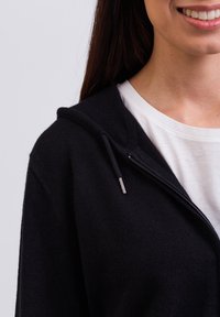 Schwarze Zip-Hoodie aus weichem Stoff mit Kapuze und silbernem Zubehör, über einem weißen T-Shirt getragen, das ein einfaches Design zeigt.