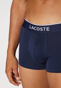 Mornarsko modri bombažni bokserji z elastičnim pasom blagovne znamke Lacoste, ki nudijo tesno prileganje in gladko teksturo. Poudarki vključujejo kontrastni bel logotip.