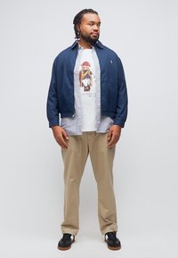 Άνδρας που στέκεται με ναυτικό μπουφάν, ανοιχτό γαλάζιο πουκάμισο, λευκό μπλουζάκι με στάμπα αρκούδας, μπεζ παντελόνια και μαύρα αθλητικά παπούτσια, κοιτάζοντας προς τα δεξιά.