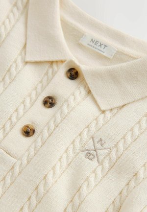 Kreemikas värvi polo-kampsun tekstuurse kaabli-silmusmustriga, kolm pruuni nööpi ja peent tikandit kangal.