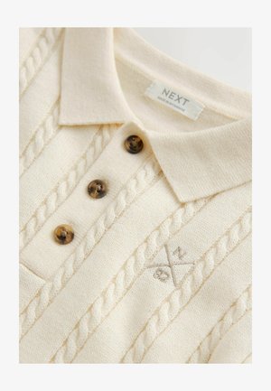 Maglione polo color crema con motivo a trecce lavorato a maglia texturizzata, tre bottoni marroni e sottili dettagli ricamati sul tessuto.