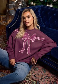 Młoda kobieta z blond włosami siedzi na wzorzystym dywanie, ubrana w fioletowy sweter z dużym różowym wzorem w kształcie kokardy, opierając się o niebieską welurową sofę.