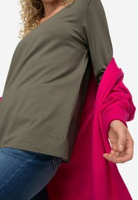Oliwkowy bawełniany top z dekoltem w kształcie litery V i krótkimi rękawami, z subtelnym logo nieskończoności. W połączeniu z jasnoróżowym kardiganem.
