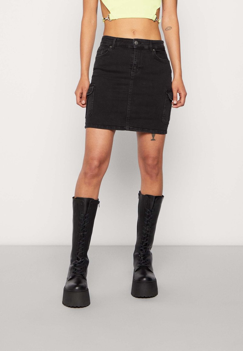 Femme portant une mini-jupe en jean noire avec des poches latérales, associée à des bottes à plateforme noires à lacets et un top court jaune.