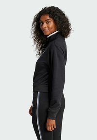 Svart sporttröja med ståkrage och ribbade muddarna, bärs med svarta byxor som har en sidostrecksdetalj. Texturerad tyg.